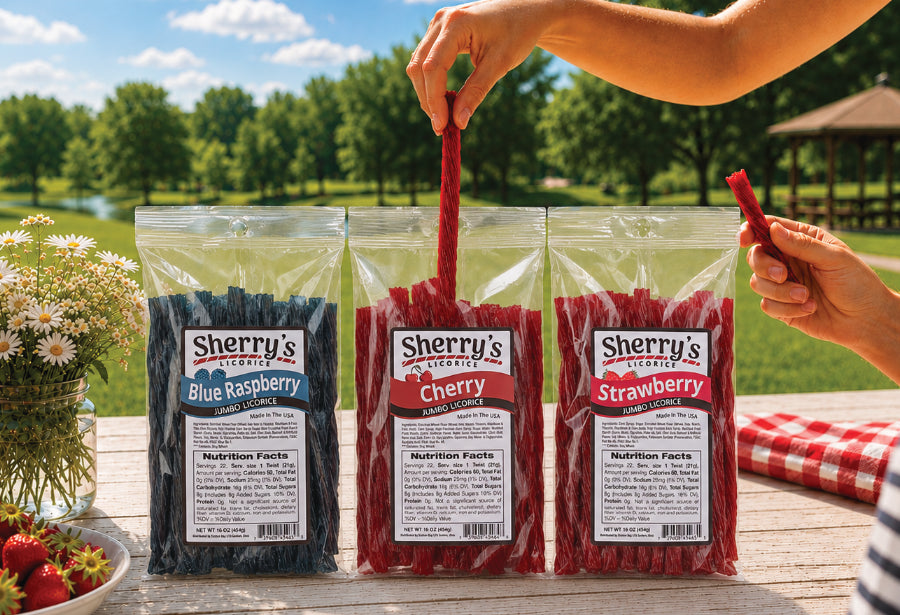 Three bags of Sherry's Licorice on a wooden table outdoors, with a person's hands holding a strawberry candy stick above along side blue raspberry and cherry bags.