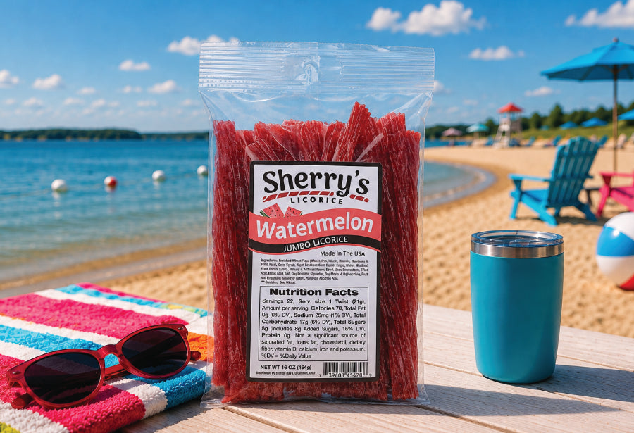 A bag of Sherry's Licorice Watermelon jumbo licorice stands on a beach table, with a blue tumbler, sunglasses, and a striped towel nearby. 